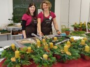 Frauen in Schürzen zeigen Adventsgestecke aus Tannenzweigen mit Bienenwachskerzen