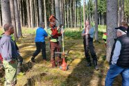 Eine Männergruppe steht im Wald, in der Mitte zeigt ein Mann ein Arbeitsgerät.