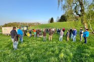 Mann spricht zu Personengruppe auf grüner Wiese, ein Traktor steht im Hintergrund.